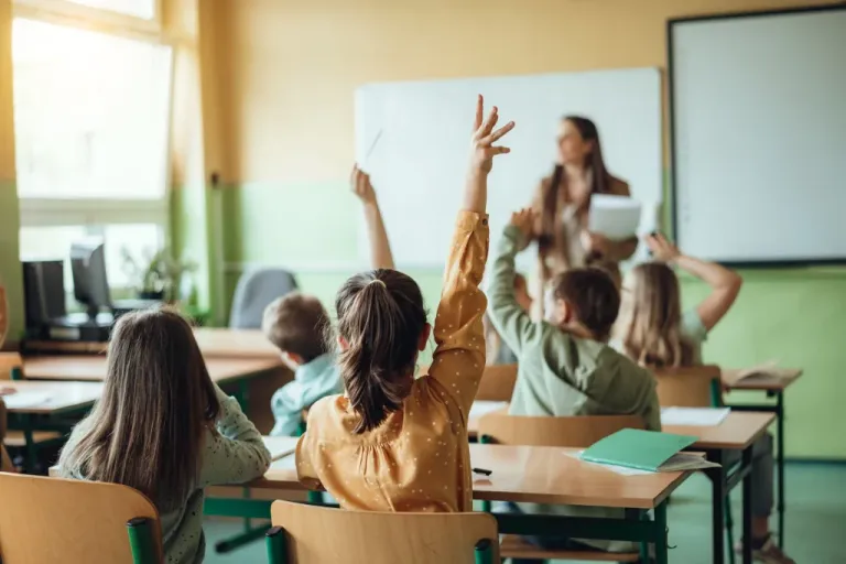 Students raising hands while a teacher asks them questions in a classroom.