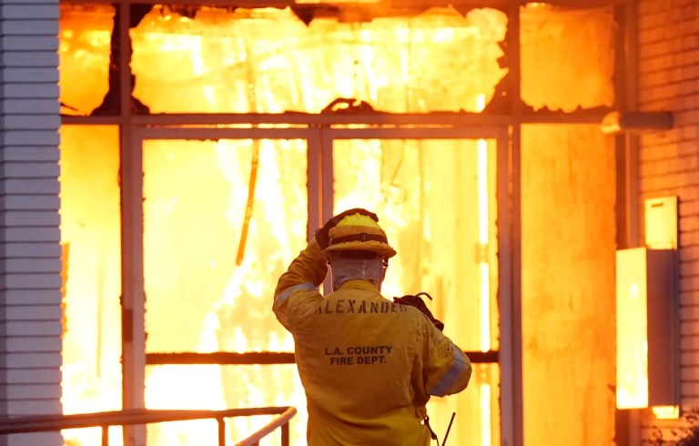 A firefighter adjusts his helmet as he looks at a Bank of America building on Lake Avenue engulfed in flames during the Eaton Fire, Wednesday, Jan. 8, 2025, in Altadena, California.