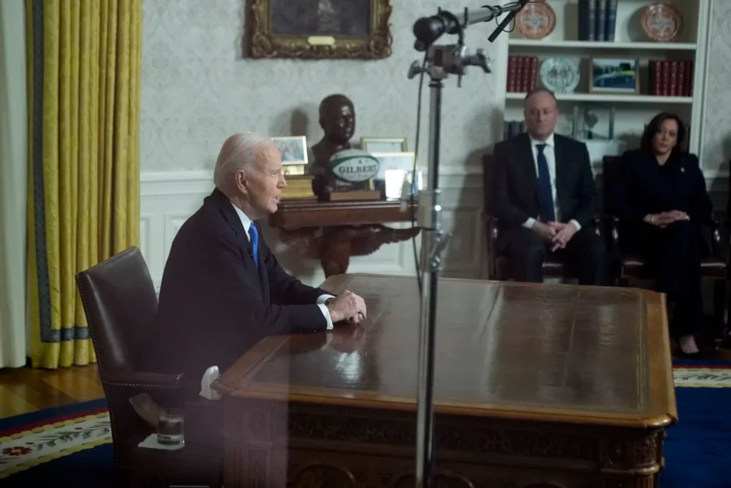 As seen through a window from the Colonnade outside the Oval Office, President Joe Biden speaks during his farewell address at the White House in Washington, Wednesday, Jan. 15, 2025, as second gentleman Doug Emhoff and Vice President Kamala Harris listen.