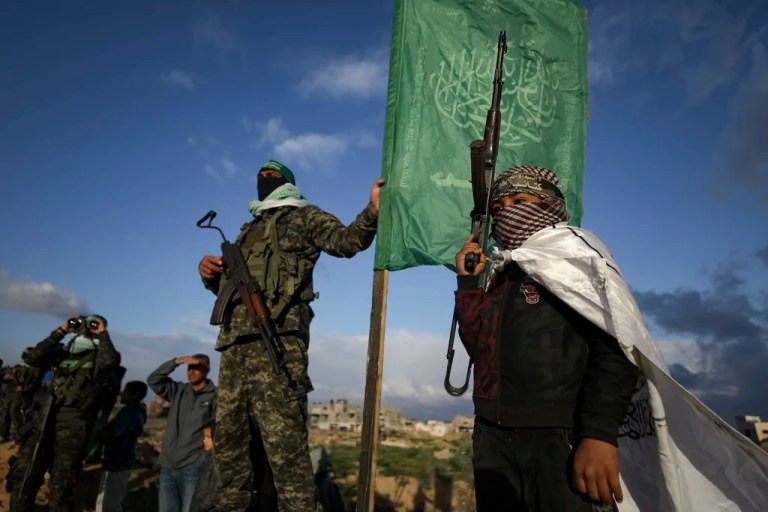 A boy holds a weapon as he stands next to a Hamas fighter standing in position ahead of handing over four bodies to the Red Cross in Khan Younis, southern Gaza Strip, Thursday, Feb. 20, 2025.