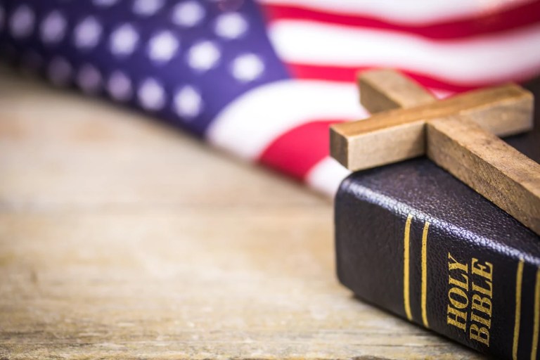 A wooden Christian cross laying on a holy Bible with an American flag background. (