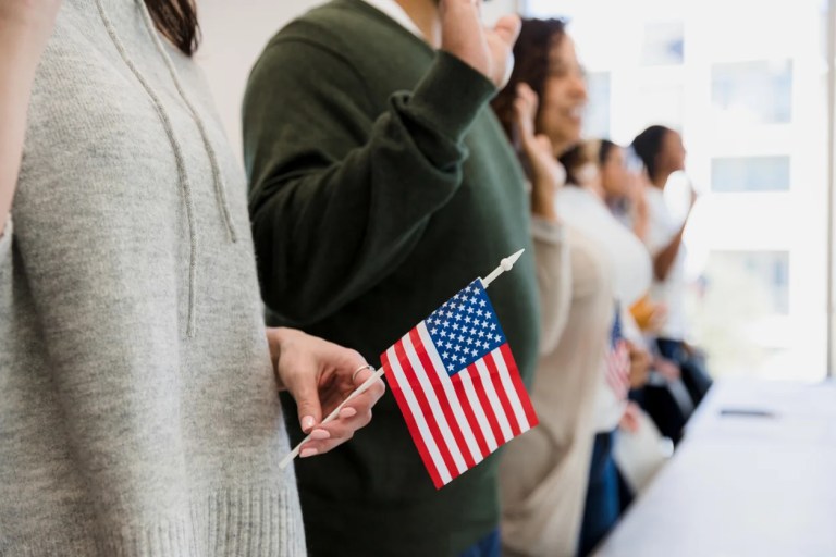 A group of multiracial people stand and raise their hand to say the pledge.