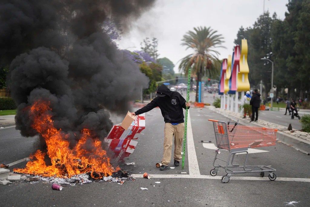 A protester places debris in a fire as Border Patrol personnel in riot gear and gas masks stand guard outside an industrial park in Paramount, Calif., on Saturday, June 7, 2025.