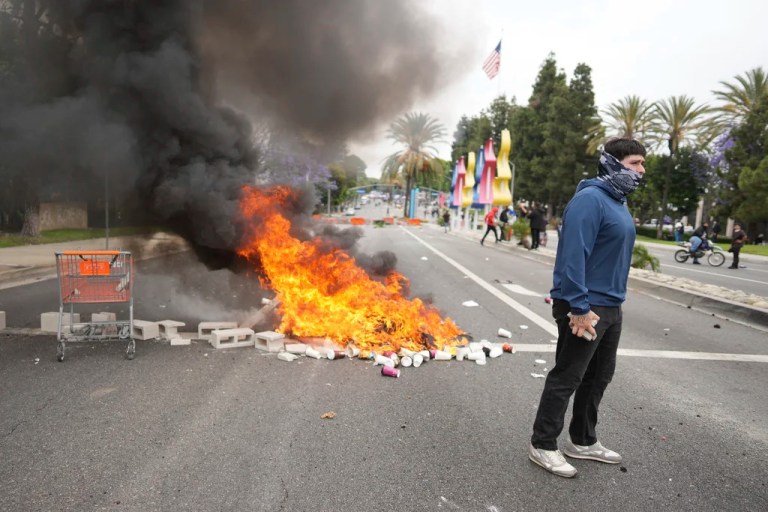 A fire burns as a protester stands across the way from Border Patrol personnel in riot gear and gas masks outside an industrial park in Paramount, California, on Saturday, June 7, 2025.