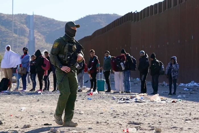 A member of U.S. Customs and Border Protection walks past a group of migrants hundreds of migrants gather along the border Tuesday, Dec. 5, 2023, in Lukeville, Ariz. The U.S. Border Patrol says it is overwhelmed by a shift in human smuggling routes, with hundreds of migrants from faraway countries like Senegal, Bangladesh and China being dropped in the remote desert area in Arizona. (AP Photo/Ross D. Franklin)