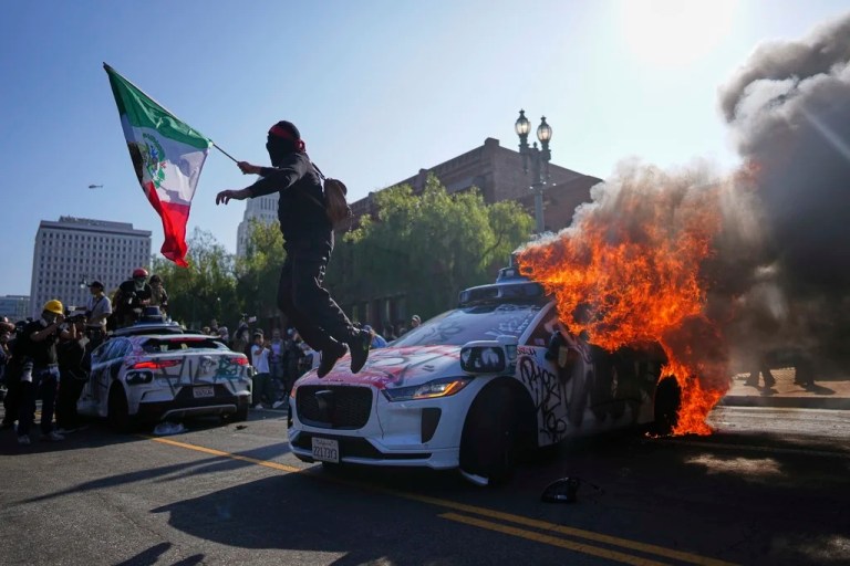 A protester leaps off a burning Waymo taxi near the Metropolitan Detention Center in downtown Los Angeles, Sunday, June 8, 2025, following last night's immigration raid protest.