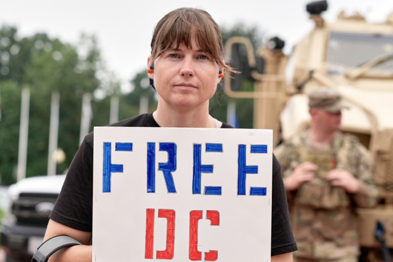 A demonstrator stands outside of Union Station as National Guard soldiers keeps watch travelers arrive at the entrance to Union Station Wednesday, Aug 20, 2025, in Washington. (AP Photo/Mariam Zuhaib)