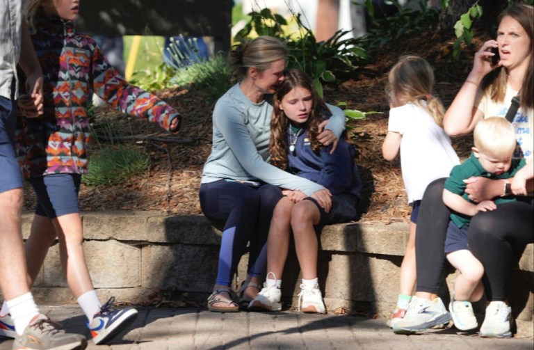A parent hugs their child outside a school as other families sit nearby.