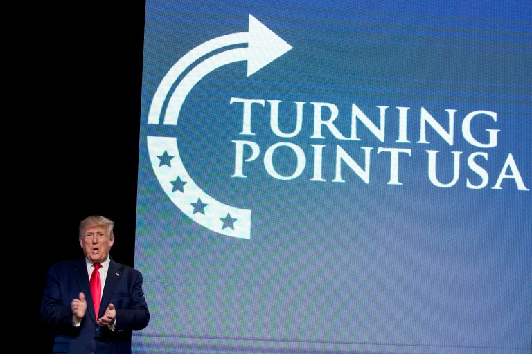 President Donald Trump clapping while standing next to a Turning Point USA screen