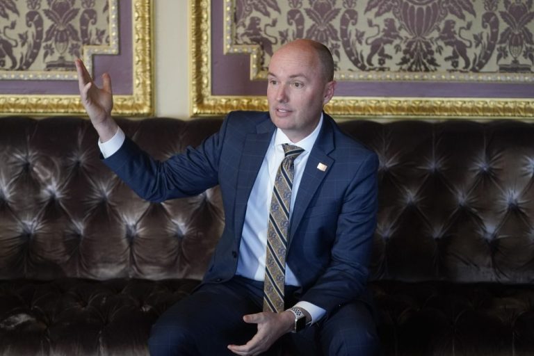 Governor Spencer Cox sitting on a brown couch in a blue suit holding up his hand