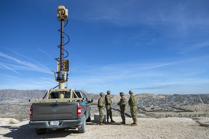 Army soldiers look at the border wall next to a surveillance vehicle.