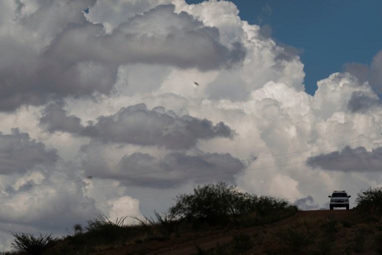 A U.S. Border Patrol vehicle travels along a trail beneath storm clouds near the U.S.-Mexico border fence.