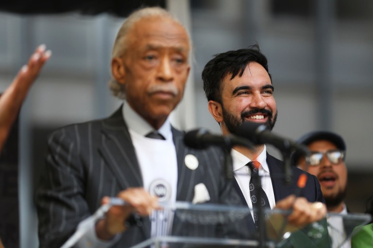 Zohran Mamdani stands and smiles behind Al Sharpton at a rally.