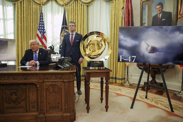 President Donald Trump and Secretary of War Pete Hegseth listen as an image of an F-47 sixth-generation fighter jet is displayed during an event in the Oval Office.