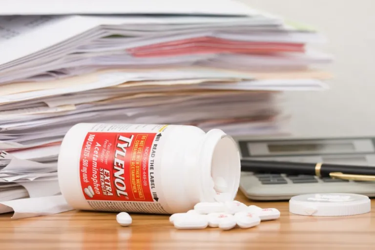 A tipped over and spilled bottle of Extra Strength Tylenol Acetaminophen Caplets in front of a pile of paperwork, a calculator, a pen, and the bottle lid. The Tylenol bottle contains 100 caplets, 500 mg each and is manufactured By: McNeil Healthcare LLC, Las Piedras, Puerto Rico For: McNeil Consumer Healthcare, Division of McNeil - PPC, Inc. Fort Washington, PA, USA