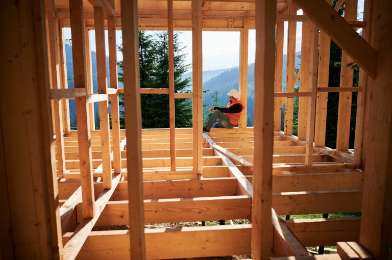 Carpenter constructing wooden frame house near the forest. Bearded man in glasses holding hammer, dressed in protective helmet and orange safety vest. Concept of ecological modern construction.