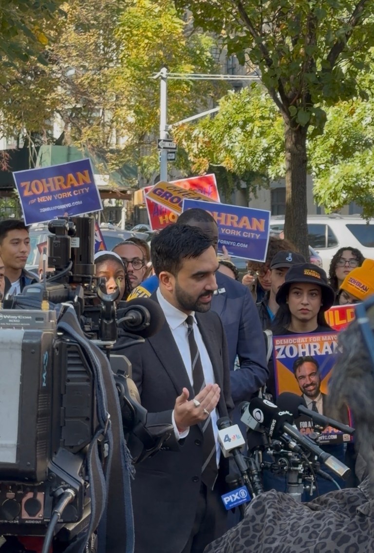Zohran Mamdani speaks to the press with supporters behind him.