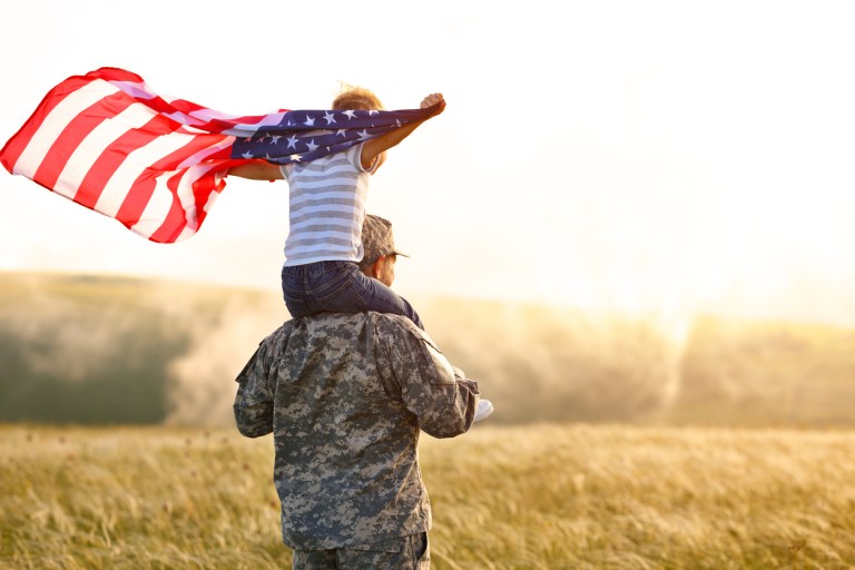 A military man carries his son, who is holding an American flag, on his shoulders in a sunny field.