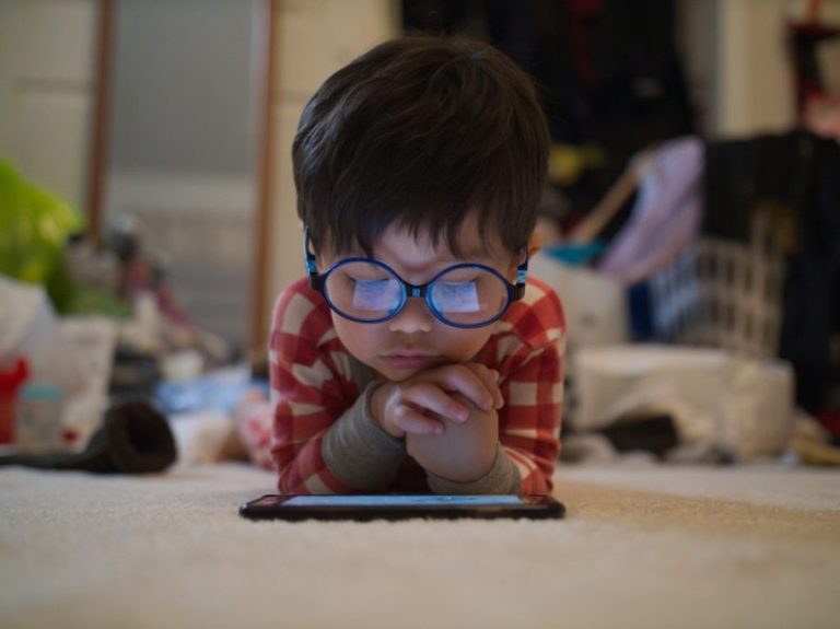 Young boy with glasses lounging on floor watching cartoons on a smartphone