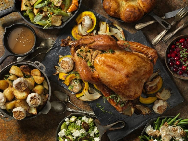 Golden roasted turkey with stuffing surrounded by traditional holiday sides, including roasted potatoes, cranberries, salad, and gravy on a rustic table.