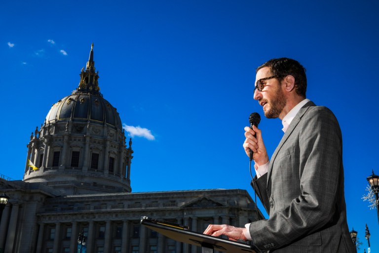 California Democratic state Sen. Scott Wiener speaks into a microphone with the San Francisco's City Hall in the background.