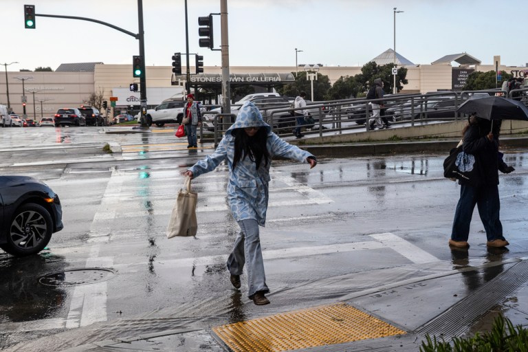 A pedestrian leaps over a stream of water while crossing 19th Avenue during a storm break in San Francisco, Friday, Dec. 26, 2025.