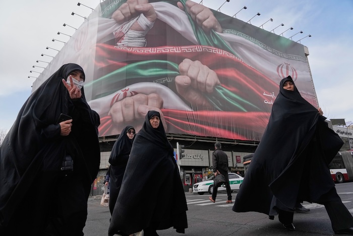 Women cross a street under a huge banner showing hands firmly holding Iranian flags as a sign of patriotism, as one of them flashes the victory sign.