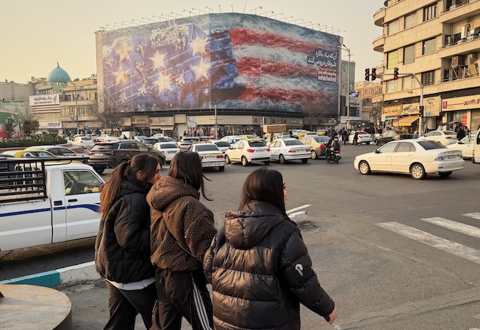 People walk in front of a billboard with a graphic showing a U.S aircraft carrier with damaged fighter jets on its deck, and sign reading in Farsi and English: 