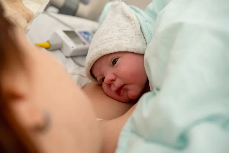 Newborn baby wearing a cozy hat, resting on a mother's chest in a hospital room.
