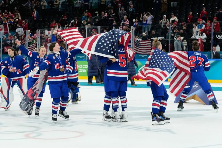 United States' Megan Keller (5), covered in the U.S. flag, gets a hug from a teammate after the United States' women's ice hockey team stand after being presented with the gold medals at the 2026 Winter Olympics, in Milan, Italy, Thursday, Feb. 19, 2026. (AP Photo/Petr David Josek)