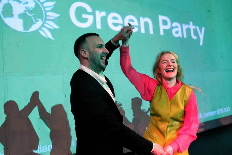 Green Party candidate Hannah Spencer and party leader Zack Polanski celebrate in front of a screen displaying the Green Party logo.