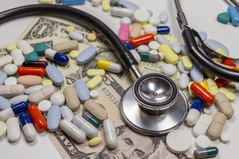 Drugs and a stethoscope sit atop bills to illustrate rising healthcare costs.