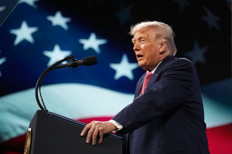 Donald Trump speaks into a microphone from behind a podium with an American flag backdrop behind him.